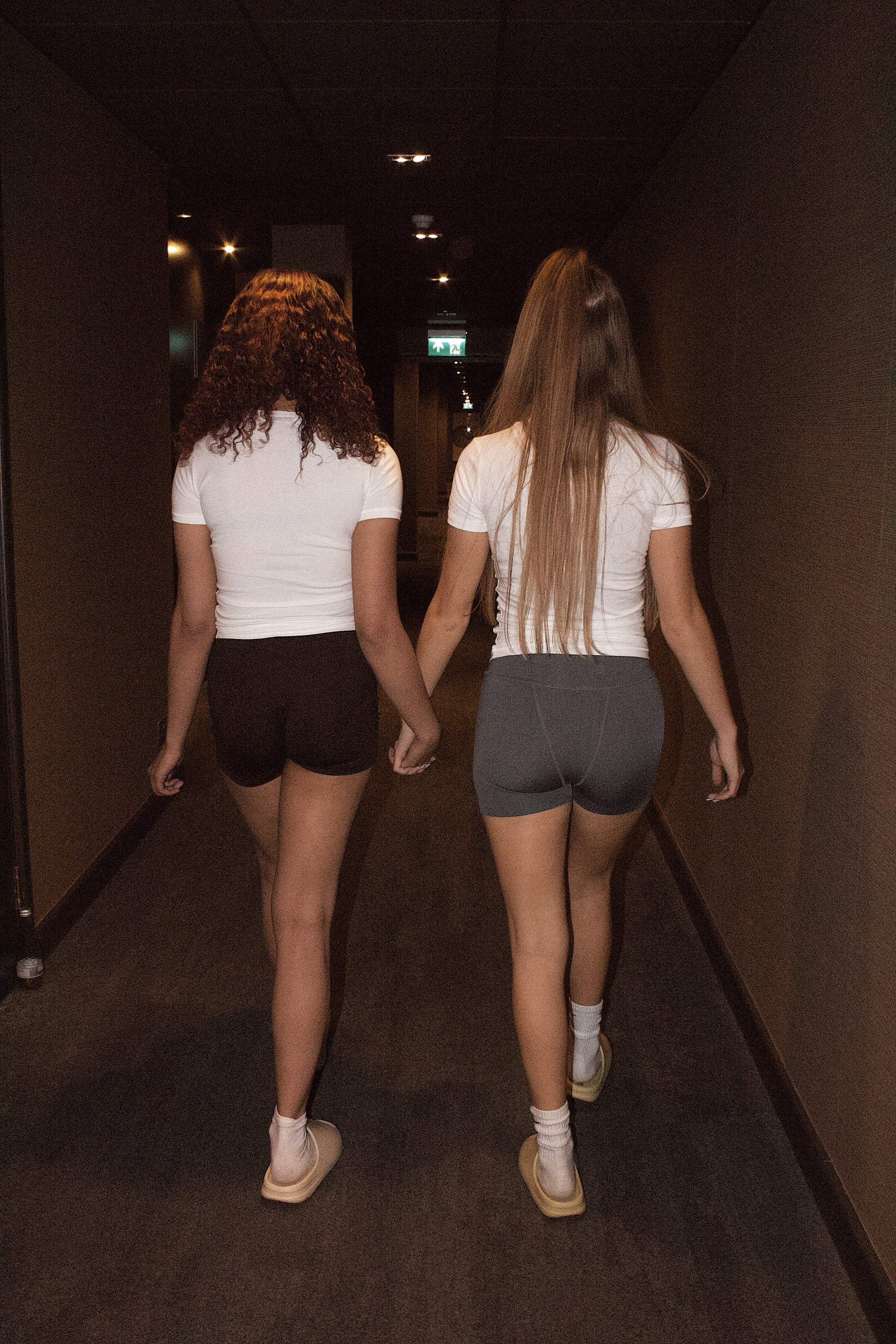 Two girls walking down a dimly lit hallway holding hands.
