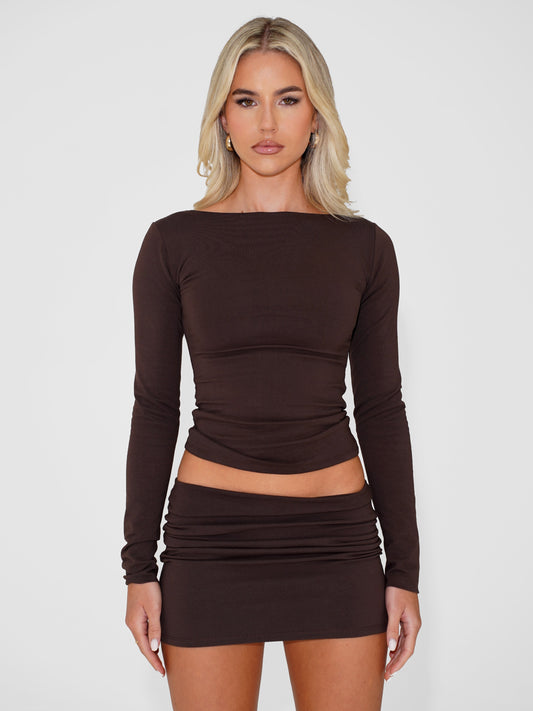 Blonde model wearing a brown long sleeve top with matching brown mini skirt, standing against a white background.
