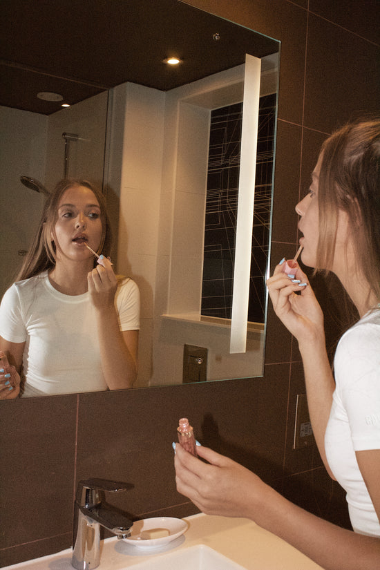 Girl applying lip gloss in front of a bathroom mirror.